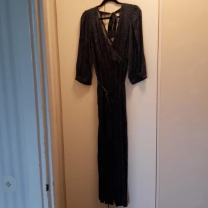 Mörkblå jumpsuit med mönster från Indiska - Blå jumpsuit från Indiska med paisleymönster. 3/4-ärmar, knytning i nacken och midjan. Resår i midjan. Mjukt, blankt, luftigt tyg som passar perfekt till sommaren. 72% viskos och 28% bomull. Endast provad, aldrig använd!