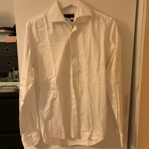 Vit slim fit skjorta från Dressmann - Klassisk vit skjorta från Dressmann i slim fit-modell. Skjortan har långa ärmar, knappar framtill och är tillverkad av bomull med lite elastan för extra komfort. Perfekt till skjorta-looken när du vill vara lite extra uppklädd.