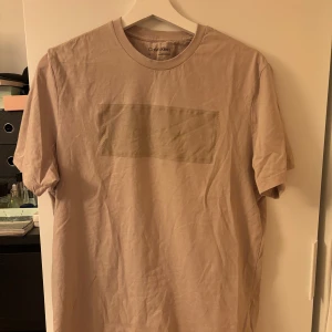 Beige t-shirt från Calvin Klein - Säljer en beige t-shirt från Calvin Klein med en diskret meshdetalj på bröstet. T-shirten har rund halsringning och är tillverkad i 100% bomull. Enkel och stilren design som passar till många outfits.