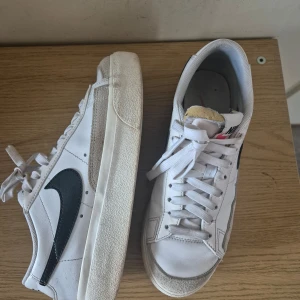 Nike Blazer Low sneakers i vitt och svart - Säljer ett par klassiska Nike Blazer Low sneakers i vitt med svart swoosh. Skorna har snörning, rund tå och detaljer i mocka framtill.