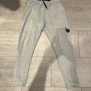 Grå mjukisbyxor från Nike - Säljer ett par grå mjukisbyxor från Nike med svart logga på benet. Byxorna har dragsko i midjan och är tillverkade i ett mjukt material med Dri-Fit-teknologi. Perfekta för träning eller chill.