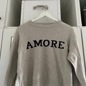 Beige sweatshirt med svart text 'AMORE' - Säljer en beige sweatshirt med svart text 'AMORE' på bröstet från Gina Tricot! Nästan aldrig använd. Perfekt för en avslappnad och trendig stil. Storlek 146/152