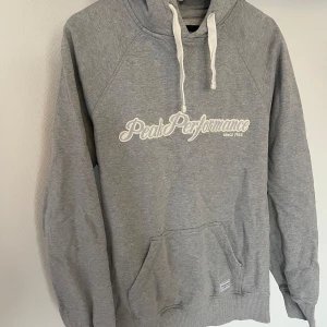 Grå hoodie från Peak Performance - Säljer en klassisk grå hoodie från Peak Performance med vit logga på bröstet och känguruficka framtill. Tröjan har huva med vita snören och ribbade muddar. Perfekt för en avslappnad stil.