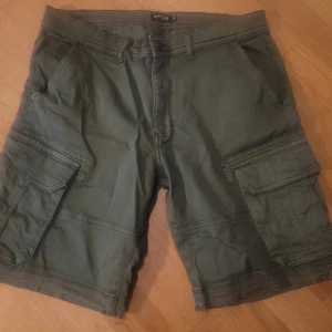 Gröna cargoshorts från On Our Terms - Säljer ett par gröna cargoshorts från On Our Terms med flera praktiska fickor på sidorna och bak. Klassisk passform med bälteshällor och knappgylf. Perfekta för en avslappnad stil. Knappt använda så bra begagnat skick. 