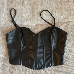 Svart korsett-topp i skinnimitation från Zara - Säljer en svart topp från Zara i skinnimitation med smala axelband och markerade sömmar framtill. Toppen har en croppad passform och är perfekt för att skapa en edgy look.