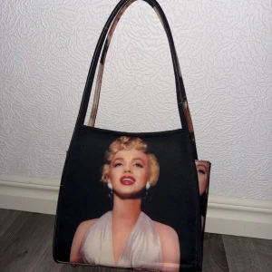 Svart axelväska med Marilyn Monroe-motiv - Cool svart axelväska med ikoniskt tryck av Marilyn Monroe på både fram- och baksidan. Väskan har rektangulär form och långa handtag, perfekt för att bära över axeln. Materialet är troligtvis syntet och trycket är färgstarkt med detaljer i vitt, rött och beige.