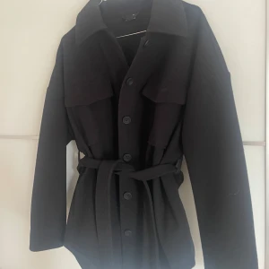Svart jacka med bälte och knappar - Säljer en svart jacka med knappar framtill och bälte i midjan. 
