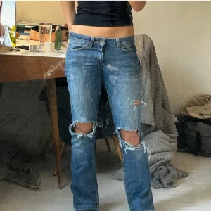 Blå bootcut jeans från Wrangler - Säljer ett par lågmidjade blå bootcut jeans från Wrangler med slitningar och hål på knäna. Jeansen har klassisk femficksdesign, låg midja och råa kanter vid hålen.