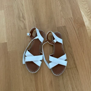 Vita sandaler med korsade remmar - Säljer ett par vita sandaler med korsade remmar och spänne vid ankeln. Skorna har platt sula och dekorativa sömmar runt sulkanten. Perfekta för varma dagar. Storlek 38