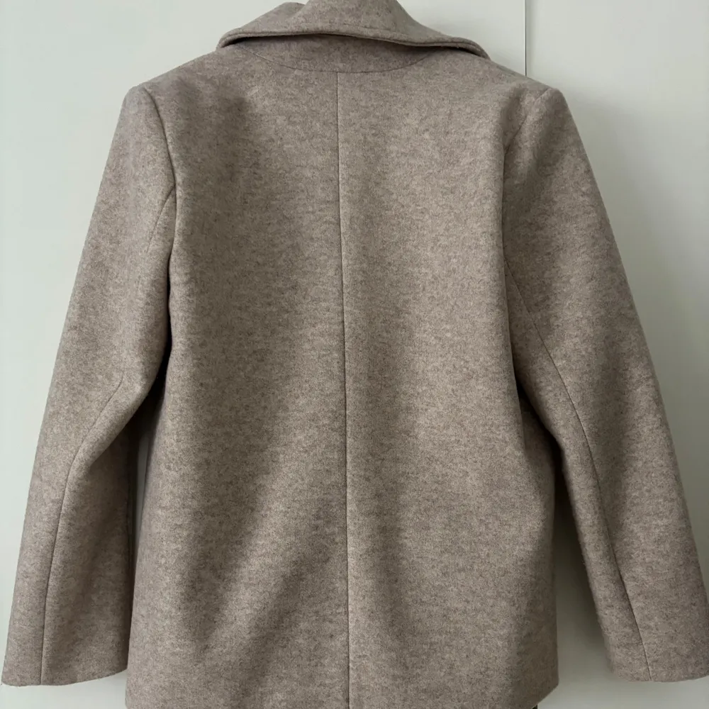 En beige kappa med ett flertal knappar framtill för stängning 🩷 (kappan kan se lite grå ut på bilden, men är mer åt det beiga hållet i verkligheten ☺️) Finns två fickor framtill!. Takit.