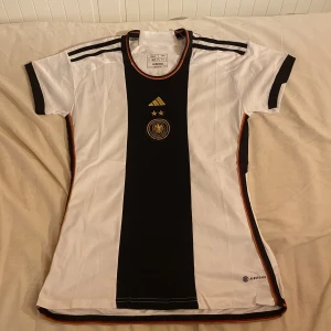 Tysklands fotbollströja från Adidas - Säljer en officiell Tyskland fotbollströja från Adidas med svart mittpanel, vita sidor och svarta detaljer. Tröjan har korta ärmar, guldiga stjärnor och broderad landslagssköld på bröstet. Slim fit-modell med klassiska ränder på axlarna.