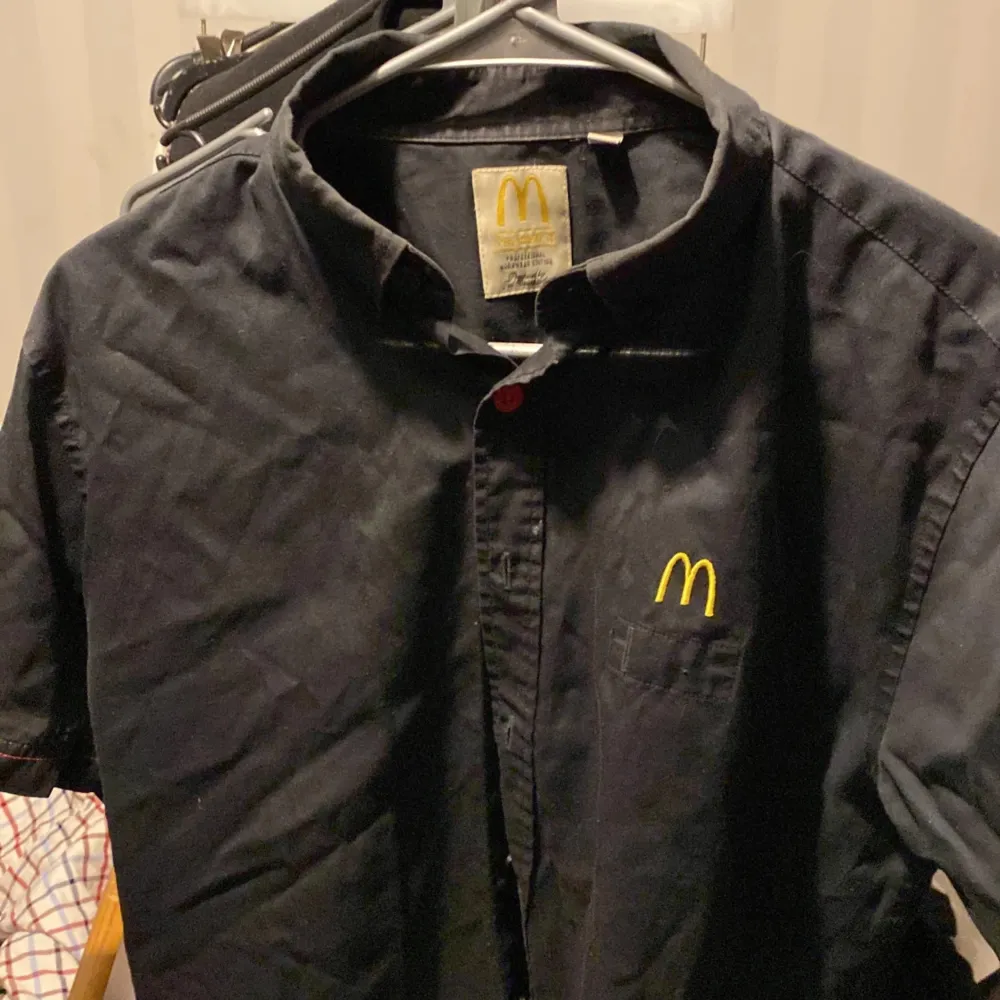Säljer en svart skjorta med kort ärm och klassisk krage. Skjortan har en gul broderad McDonald's-logga på bröstfickan och knäppning framtill. Perfekt för dig som vill sticka ut eller gillar unika plagg!. Paidat.