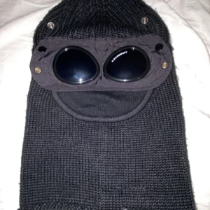 Svart stickad balaclava med glasögon från C.P. Company - Säljer en svart stickad balaclava från C.P. Company med inbyggda glasögon. Balaclavan täcker hela huvudet och har en unik design med runda linser framtill. Perfekt för kalla dagar och streetwear-stil.