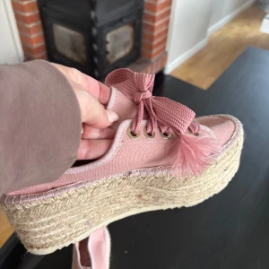 Rosa sneakers med platåsula och tofsar - Säljer ett par rosa sneakers med breda snören och dekorativa tofsar framtill. Skorna har en hög platåsula i flätad jute som ger en espadrillos-känsla. Perfekta för dig som vill sticka ut med dina skor!