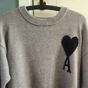 Grå stickad tröja från Ami - Säljer en grå stickad tröja från Ami med svart hjärta och bokstaven A broderat på bröstet. Tröjan har rund halsringning och långa ärmar. Perfekt för dig som gillar stilrena detaljer. Aldrig använd. Kvitto saknas.