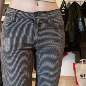 Randiga jeansbyxor från Cheap Monday - Säljer ett par lådmidjade grå/bruna jeansbyxor med vita smala ränder från Cheap Monday. Byxorna har slim passform, klassisk femficksdesign och stängs med dragkedja och knapp. Perfekta för dig som gillar en retrostil med edge.