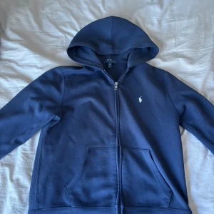 Marinblå hoodie från Polo Ralph Lauren - Säljer en marinblå hoodie från Polo Ralph Lauren med dragkedja och huva. Klassisk design med vit broderad logga på bröstet och två fickor framtill. Perfekt för en avslappnad stil.