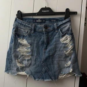 Blå jeanskjol från Hollister - Säljer en blå jeanskjol från Hollister med slitna detaljer och rå kant nertill. Kjolen har klassisk femficksdesign och knappgylf. Broderade detaljer på bakfickorna. Perfekt för dig som gillar en tuff och avslappnad stil.