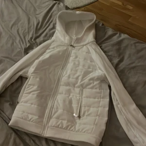 Vit quiltad hoodiejacka med dragkedja - Snygg vit hoodiejacka med quiltade partier framtill och på sidorna. Jackan har huva, hel dragkedja och två fickor med dragkedja. Perfekt för lager-på-lager och en stilren look.