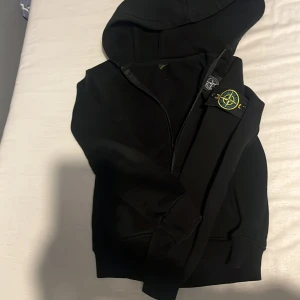 Svart hoodie från Stone Island - Säljer en svart hoodie från Stone Island med klassisk patch på ärmen och logga på insidan. Tröjan har huva och ribbade muddar vid ärmslut och nederkant. Perfekt för dig som gillar streetwear och vill ha något stilrent.