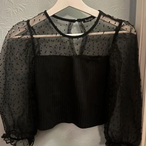 Svart transparent blus med prickigt mönster från Boohoo - Säljer en svart blus från Boohoo med transparenta, puffiga ärmar och små prickar. Blusen har rund halsringning, knäppning bak i nacken och croppad passform. Perfekt för dig som gillar trendiga detaljer och volym i ärmarna.