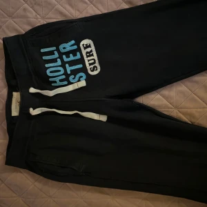 Svarta mjukisbyxor från Hollister - Svarta mjukisbyxor från Hollister med blå och vit text på ena benet. Byxorna har dragsko i midjan och är perfekta för chill dagar. Snygg och avslappnad stil med tydlig logga. Knappt använda 🩵