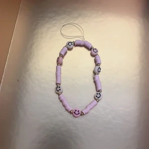 Lila pärlarmband med smileys - Ett färgglatt armband med lila pärlor och vita pärlor med svarta smiley-ansikten. Armbandet har elastisk tråd och passar perfekt som accessoar för att liva upp din stil.
