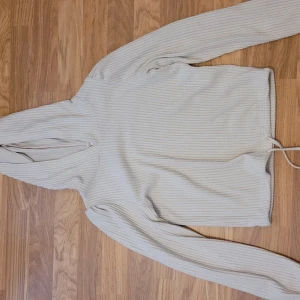 Beige ribbad hoodie - Mysig beige hoodie med ribbad struktur och långa ärmar. Tröjan har huva och dragsko nertill för en avslappnad look. Perfekt för chill dagar eller när du vill vara extra bekväm.