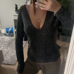 Svart glittrig v-ringad tröja - Säljer en svart långärmad tröja med glittrigt material och djup v-ringning. Perfekt för dig som vill sticka ut lite extra. Snygg passform och skön att bära.
