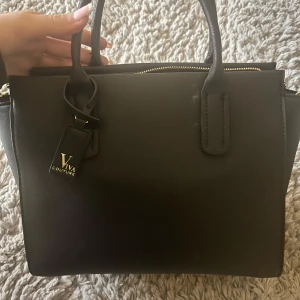 Svart handväska från Viva Couture - Snygg svart handväska från Viva Couture med två handtag och guldfärgad dragkedja. Väskan har en stilren och klassisk design, perfekt för att bära dina viktigaste saker. Tillverkad i syntetmaterial.