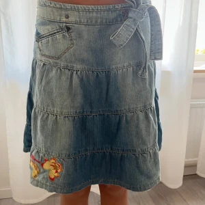 Jeanskjol med  rosett  - Säljer en blå jeanskjol med volanger och broderade blommor i gult och rött. Kjolen har knytband i midjan och en liten ficka framtill. Perfekt för dig som gillar en avslappnad och söt stil.