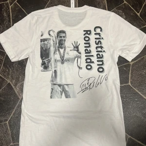 Vit Cristiano Ronaldo t-shirt med tryck stl: 158-164 - Vit t-shirt med stort svartvitt tryck av Cristiano Ronaldo som firar med en pokal. Texten 'Cristiano Ronaldo' och en tryckt autograf på framsidan. På bröstet finns CR7-logga och en get i guld. Rund halsringning. Helt ny också oanvänd. Den är gjord av bomull. 