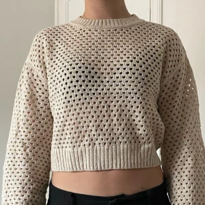 Beige croppad stickad tröja - Säljer en beige croppad stickad tröja med hålmönster och ribbade muddar. Tröjan har lång ärm och rund halsringning, perfekt för lager-på-lager och en chill look.