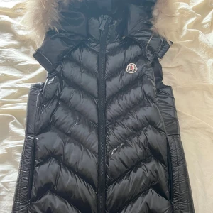 Svart dunväst från Moncler med päls - Säljer en svart glansig dunväst från Moncler med dragkedja och huva med pälskant. Västen har Moncler-logga på bröstet och ett klassiskt quiltat mönster. Perfekt för lager-på-lager under kyliga dagar. Pris går att diskutera 