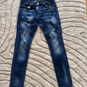 Mörkblå Dsquared2 ICON jeans med slitningar - Säljer ett par mörkblå Dsquared2 ICON jeans med tydliga slitningar och distressed detaljer framtill. Klassisk femficksmodell med smal passform och ikonisk svartvit patch bak 