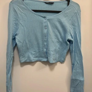 Ljusblå croppad ribbad topp - Säljer en ljusblå croppad topp med långa ärmar och knappar framtill. Toppen har ribbad struktur och en enkel, stilren design som passar perfekt till jeans eller kjol.
