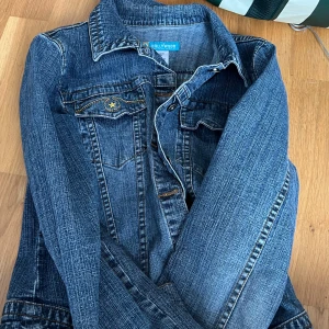 Blå jeansjacka från Hollywood - Säljer en klassisk blå jeansjacka från Hollywood med snygga sömmar och bröstfickor med knappdetalj. Jackan har krage och knäppning framtill. Perfekt att slänga över en t-shirt eller hoodie för en cool look.