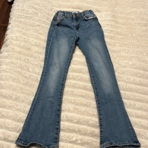 Blå bootcut jeans, storlek 140 - Säljer ett par blå bootcut jeans med papperspåse-midja och klassiska fickor både fram och bak. Jeansen har en snygg tvätt och är perfekta för dig som gillar en trendig och avslappnad stil.