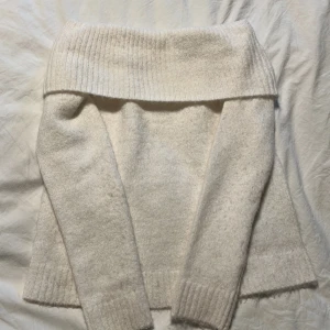 Offshoulder stickad tröja i cremevit - Mysig cremevit stickad tröja med bred offshoulder-krage och långa ärmar. Tröjan har ribbade muddar vid ärmslut och nederkant, perfekt för lager på lager under kyliga dagar.