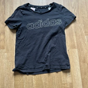Svart t-shirt från Adidas - Svart t-shirt från Adidas med stor vit logga framtill. Klassisk passform med rund halsringning och korta ärmar. Perfekt för en sportig och avslappnad stil.