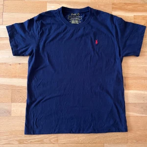 Mörkblå t-shirt från Polo Ralph Lauren - Säljer en mörkblå t-shirt från Polo Ralph Lauren med klassisk röd broderad logga på bröstet. T-shirten har rund halsringning och är tillverkad i mjuk bomull. Perfekt för en stilren och avslappnad look.