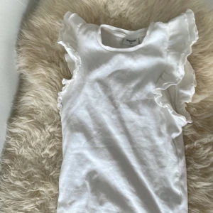 Vit volangtopp från Kappahl - Andvänd en gång. Säljer en söt vit topp från Kappahl med volangdetaljer längs ärmhålen. Toppen är ärmlös och har en rund halsringning. Perfekt för en fräsch och stilren look.