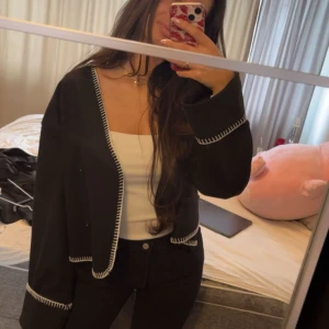 Svart kofta med vita sömmar från Gina Tricot - Säljer en svart kavaj från Gina Tricot med dekorativa vita sömmar längs kanterna och ärmsluten. Kavajen har lång ärm, knäppning framtill och en croppad passform. Perfekt för dig som vill ha en stilren men unik look. Storlek S 