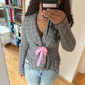 Oddmolly kofta - Säljer en svart och vit mönstrad kofta med lång ärm och en söt rosa knytdetalj i midjan. Koftan har en figurnära passform och är perfekt för att piffa upp vardagslooken.