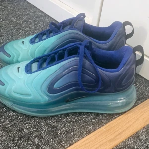 Nike Air Max 720 blå/turkos sneakers - Nike Air Max 720 sneakers i blå och turkos med futuristisk design och hög sula. Skorna har snörning och är tillverkade i syntetmaterial. Perfekta för dig som gillar streetwear och vill sticka ut med färg.