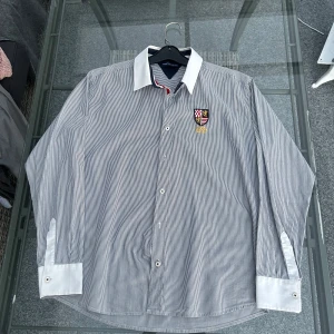 Randig skjorta från Tommy Hilfiger - Snygg blåvit randig skjorta från Tommy Hilfiger med vit krage och manschetter. Skjortan har broderad logga och emblem på bröstet samt knappar framtill. Perfekt för en stilren look. #tommyhilfiger #herrskjorta #preppystyle #klassisklook #stilren #designerskjorta #newyorkstyle #vitskjorta #randigskjorta #secondhandfashion #plickfinds #th #herrmode #skjortamedstil  #mensshirt #preppystyle #classiclook #smartcasual #designershirt #newyorkvibes #stripedshirt #whiteshirt #stylishmenswear #secondhandst