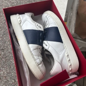 Valentino sneakers med blå detalj - Snygga vita Valentino sneakers med mörkblå rem över mitten och nitar på sulan. Skorna har snörning och är tillverkade i skinn. Stilrena och exklusiva sneakers, väl använda därav bra pris. Skokartong + extra snörning ingår💕💕