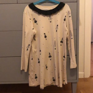 Vit klänning med svart krage från LIVLY - Supersöt vit klänning från LIVLY med svart volangkrage och lång ärm. Klänningen har ett gulligt svart mönster med små tecknade figurer över hela tyget. Perfekt för dig som gillar lekfull stil!