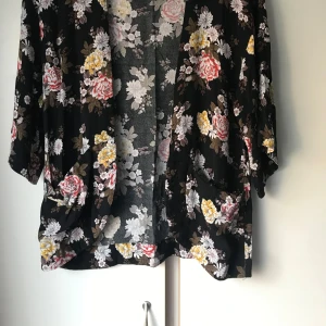Svart blommig kimono-jacka - Säljer en svart kimono-inspirerad jacka med blommigt mönster i rosa, vitt och gult. Jackan har trekvartsärmar och en lös passform, perfekt att slänga över en topp för en snygg look.