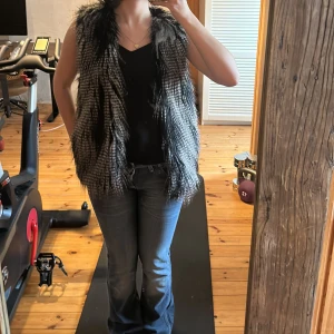 Svartvit pälsväst - Säljer en svartvit pälsväst med längre fuskpäls och öppen front. Västen har ett mönster som påminner om djurpäls och är perfekt för att ge outfiten en cool touch.